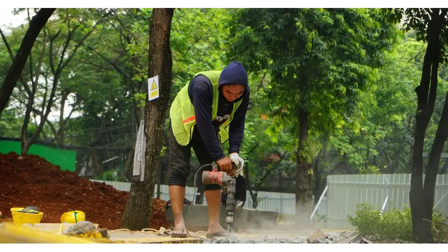A construction worker in casual attire operates a drill on a site surrounded by greenery.